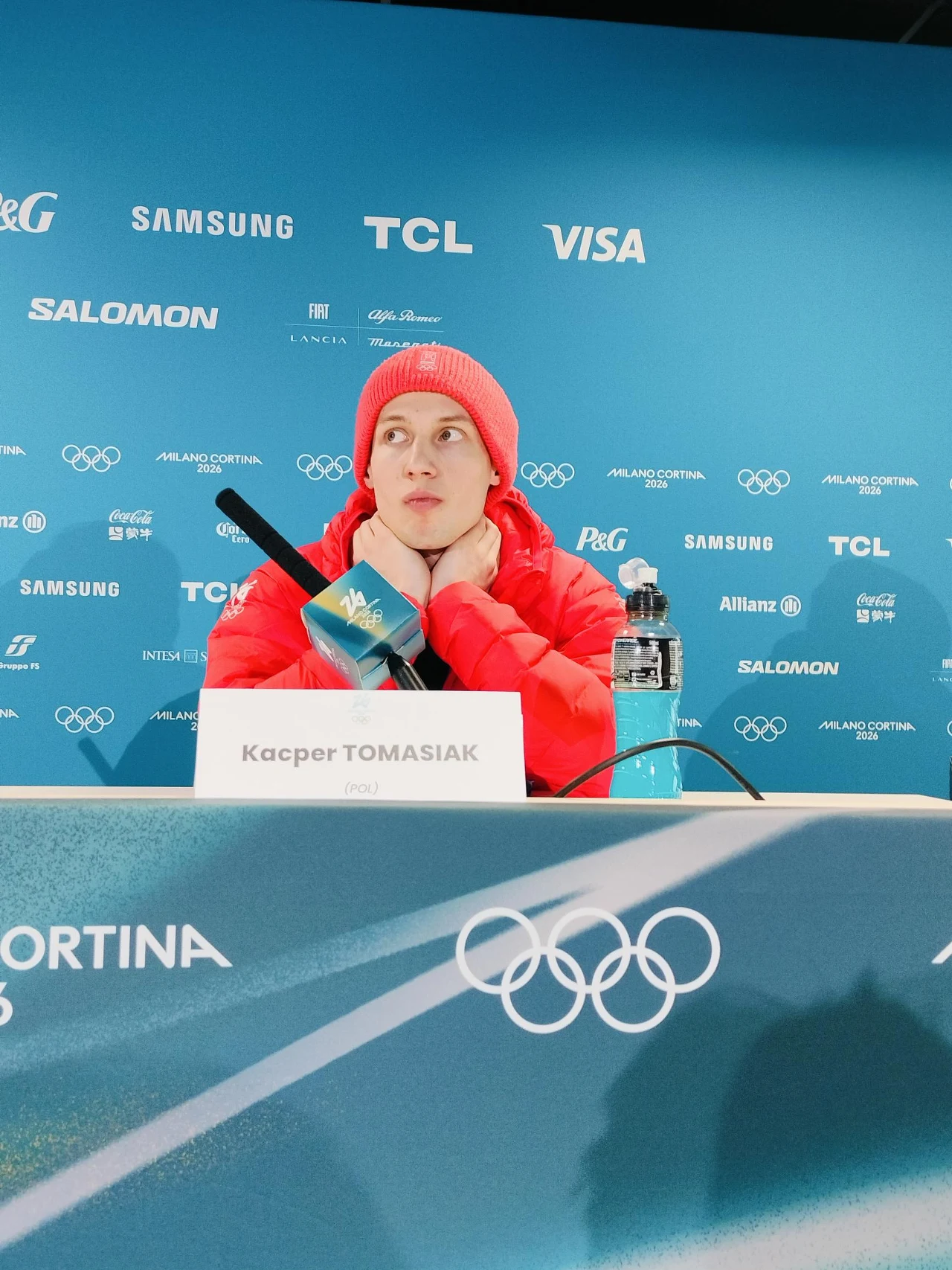 Zawodnik ubrany w czerwoną kurtkę i czapkę siedzi przy stole konferencyjnym z mikrofonem i butelką wody, przed nazwiskiem na tabliczce, na tle niebieskiej ściany oznaczonej logotypami sponsorów oraz symbolami olimpijskimi.