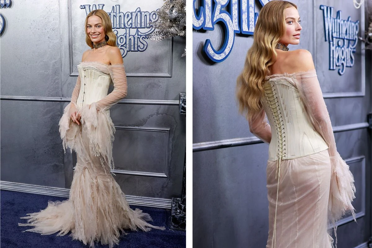 Kobieta w eleganckiej, jasnej sukni z gorsetem i długim trenem z piórami pozuje na tle ścianki promocyjnej wydarzenia, a jej włosy są rozpuszczone i ułożone w fale.