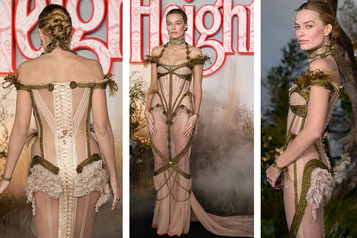 Kobieta w misternie zdobionej, jasnej sukni z przezroczystymi i sznurowanymi elementami oraz efektownymi dodatkami, stojąca na tle rozmytej grafiki i roślinności. Jej stylizacja podkreśla sylwetkę i zwraca uwagę nietuzinkowym projektem.