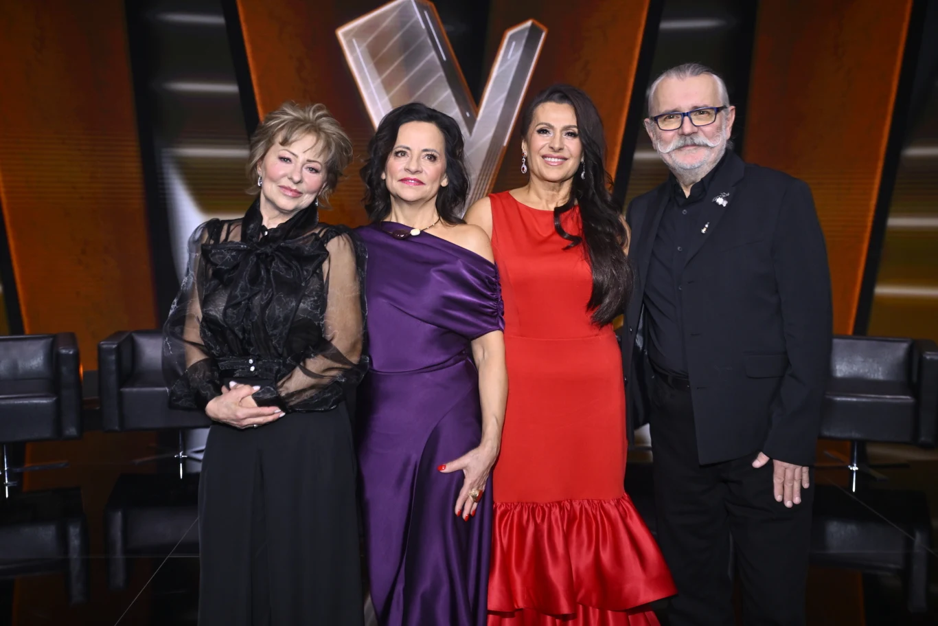 Finaliści "The Voice Senior" - od lewej: Małgorzata Skrzypczak, Grażyna Osmala, Violetta Kapcewicz i Artur Słowik Cztery dorosłe osoby ustawione razem na tle dużego, podświetlonego logotypu w kształcie litery V, trzy kobiety w eleganckich sukniach i mężczyzna w garniturze z białą brodą, wszyscy patrzą w stronę obiektywu.