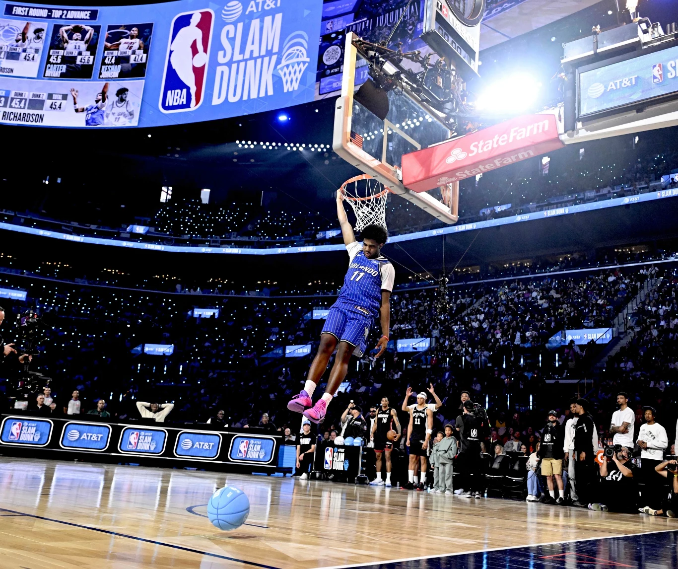 Zawodnik unoszący się w powietrzu podczas efektownego wsadu do kosza na profesjonalnej hali. Pełna publiczność oraz jaskrawe światła dodają dynamizmu i atmosfery wielkiego wydarzenia sportowego. Na boisku widać upuszczoną piłkę.