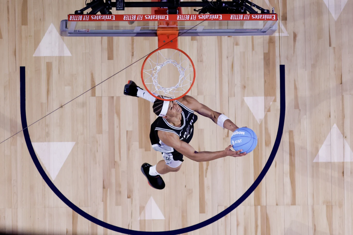 Zawodnik koszykówki w czarnej koszulce wykonuje wsad do kosza na hali sportowej, ujęcie z góry pokazuje energię i dynamikę momentu.