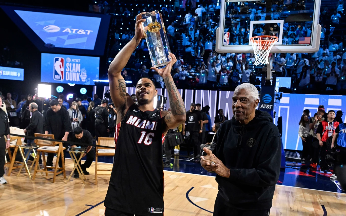 Koszykarz drużyny Miami trzyma uniesione trofeum w górze, stojąc na środku boiska, obok niego znajduje się starszy mężczyzna klaszczący w dłonie, a w tle widać publiczność oraz innych uczestników wydarzenia.