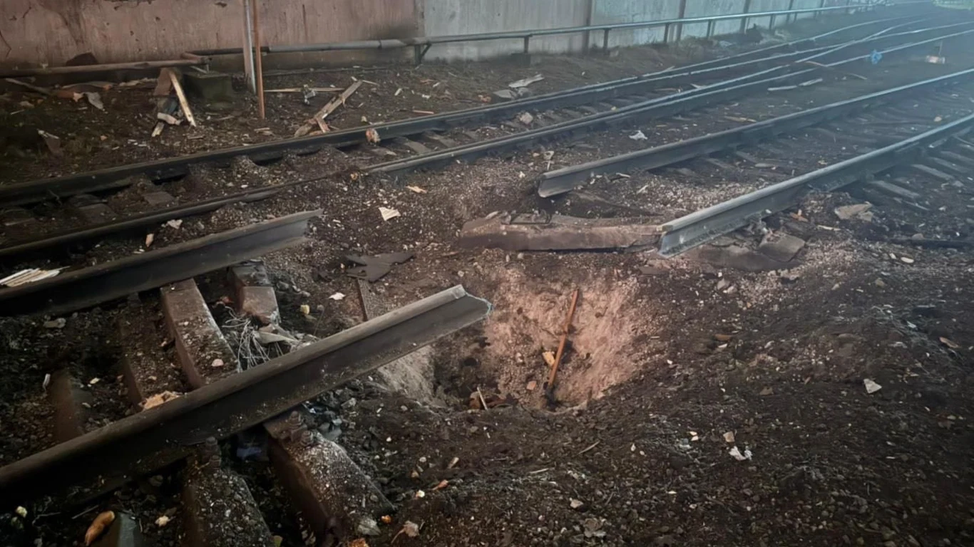 Zniszczone tory kolejowe z dużym kraterem w ziemi pomiędzy szynami, wokół rozproszone fragmenty drewna i metalu, tory wyraźnie przerwane na skutek silnego wybuchu.