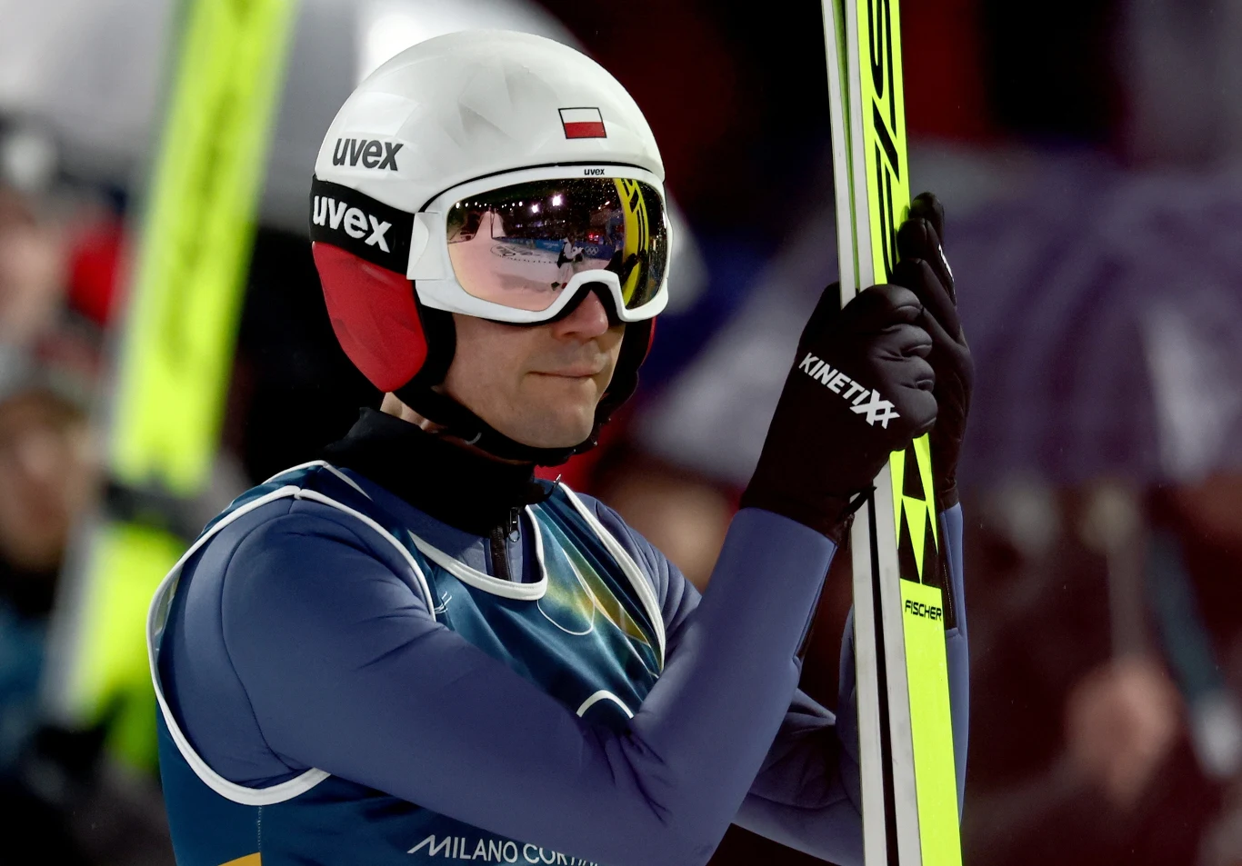 Skoczek narciarski w kasku z polską flagą na stroju sportowym, trzymający narty, w tle lekkie zamazanie tłumu i światła stadionu.