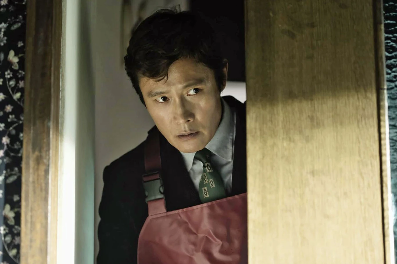 Mężczyzna w garniturze i czerwonym fartuchu wygląda zza drewnianych drzwi z niepokojem, światło podkreśla jego poważny wyraz twarzy.