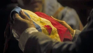 Flaga o czerwono-niebiesko-białych pasach z żółtym słońcem i gwiazdami trzymana w obu dłoniach przez osobę, w tle widoczne niewyraźne postacie, scena o nastrojowym, przyciemnionym oświetleniu.