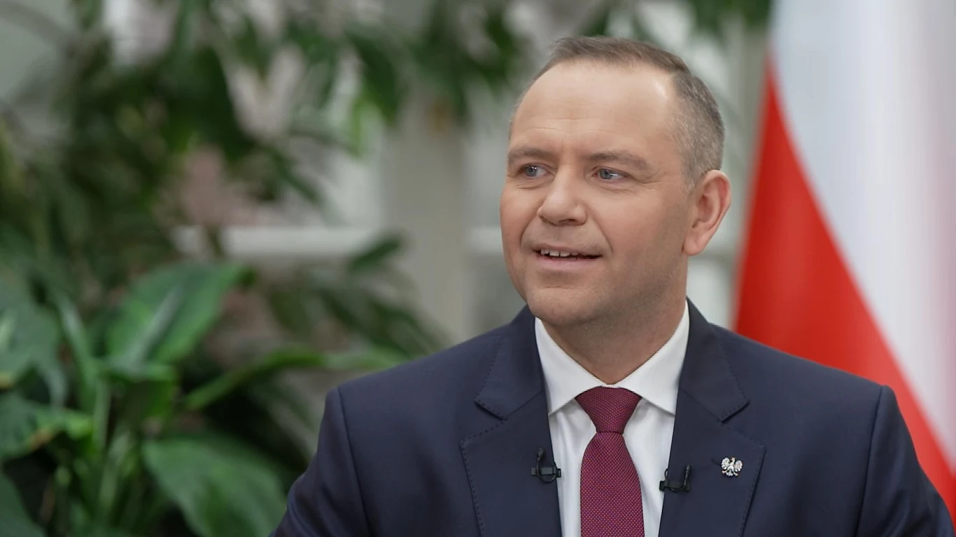 Uśmiechnięty mężczyzna w garniturze i krawacie siedzący na tle roślin oraz polskiej flagi, z przypiętym mikrofonem przy klapie marynarki.