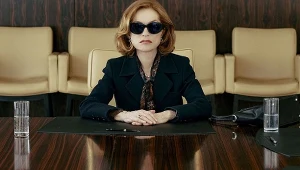 Kobieta w ciemnych okularach siedząca przy dużym, błyszczącym stole konferencyjnym, ubrana elegancko w ciemny żakiet i apaszkę, z dłońmi splecionymi na czarnych dokumentach. W tle beżowe fotele, przed nią szklanki z wodą i czarna torebka.