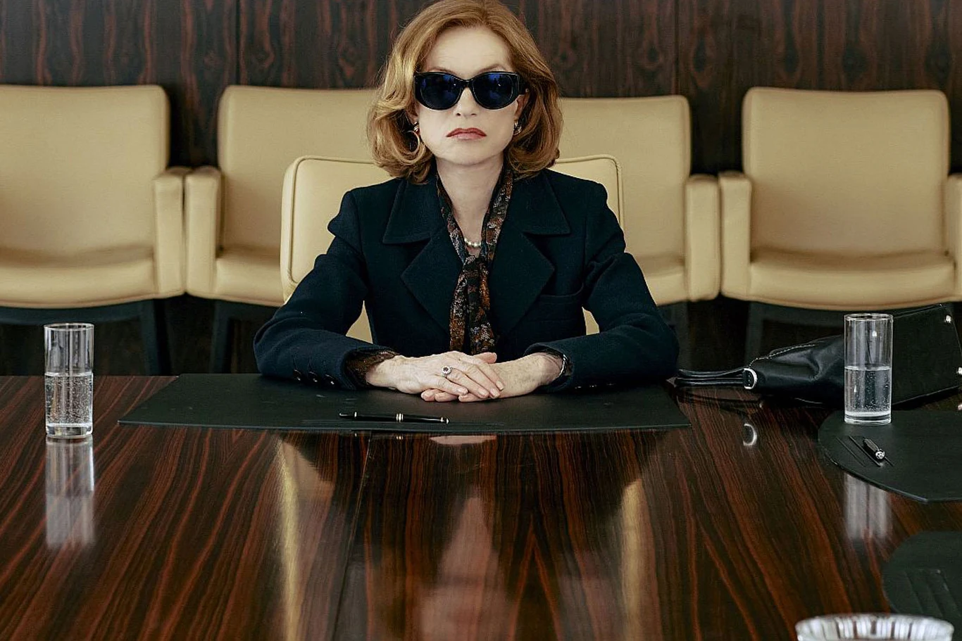 Kobieta w ciemnych okularach siedzi przy dużym, drewnianym stole konferencyjnym. Przed nią leżą dokumenty oraz długopis, po bokach dwie szklanki z wodą. Za nią znajdują się beżowe fotele na tle ciemnej, drewnianej ściany.