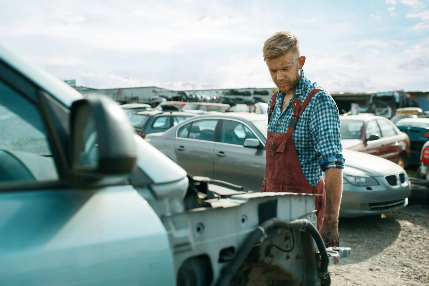 Rozbite auto nie zawsze oznacza złom. Kluczowe jest to, jak i gdzie zostało naprawione. Mężczyzna w roboczych ogrodniczkach i koszuli w kratę stoi na złomowisku samochodowym, patrząc na rozbite auto. W tle widać inne uszkodzone pojazdy ustawione na placu.