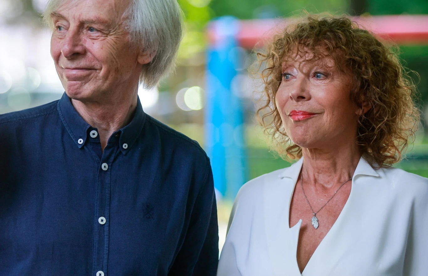 Włodzimierz Korcz, Alicja Majewska Starszy mężczyzna w granatowej koszuli i kobieta o rudych, kręconych włosach w białym żakiecie stoją razem na tle zieleni, oboje patrzą w różnych kierunkach, sprawiają wrażenie zadowolonych i spokojnych.