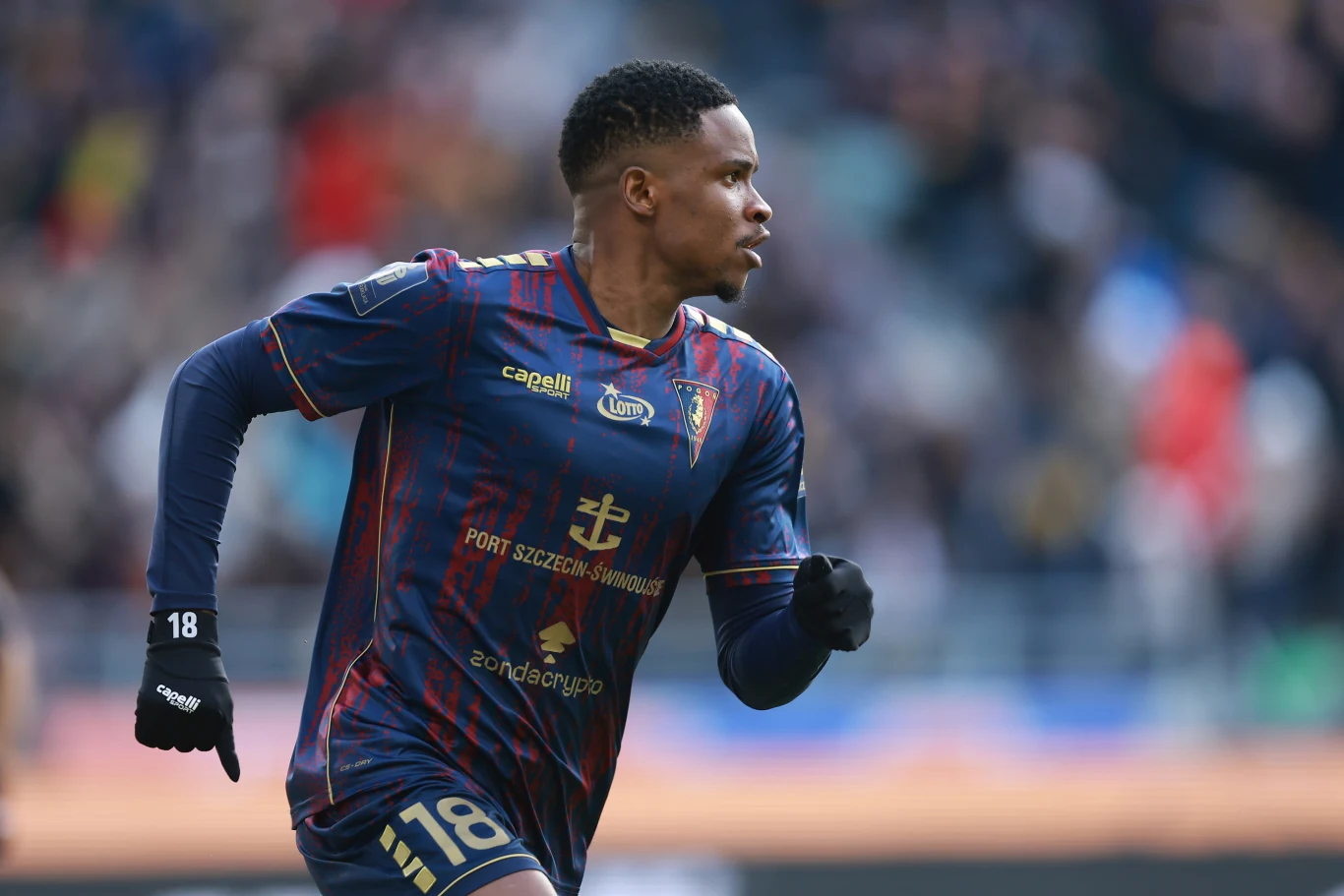 Nigeryjczyk Paul Mukairu otworzył wynik spotkania już w piątej minucie gry Piłkarz w granatowo-czerwonym stroju biegnie po murawie stadionu, skupiony na akcji podczas meczu, na trybunach widoczny rozmyty tłum kibiców.