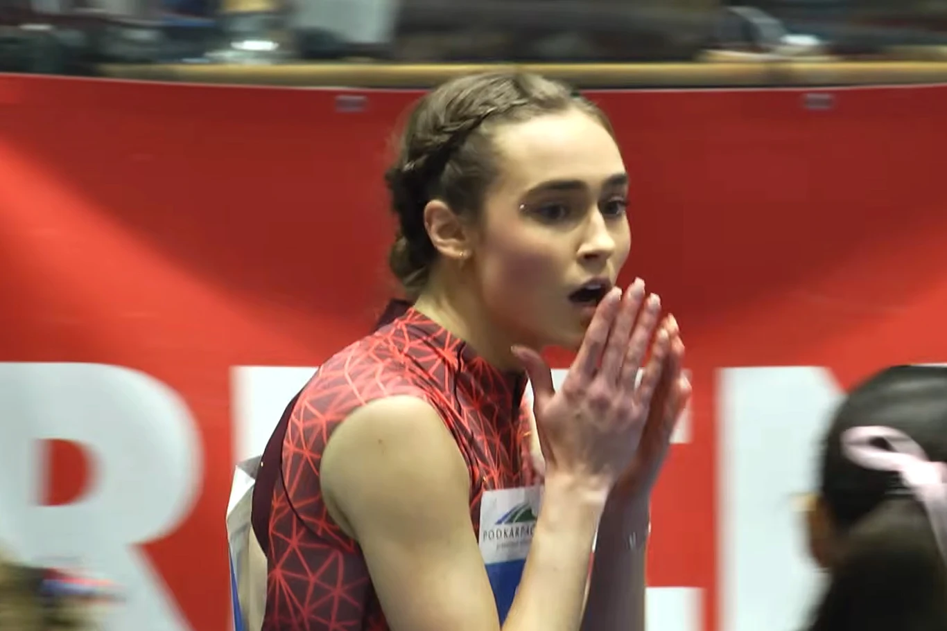 Młoda lekkoatletka w sportowym stroju stoi na tle czerwonego baneru, wyrażając emocje poprzez gest splecionych rąk przy twarzy, obok rozmyte sylwety innych uczestników.