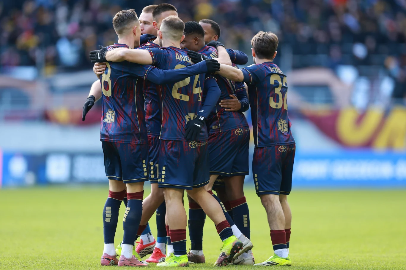 Piłkarze Pogoni przypuścili szturm na bramkę rywala od pierwszych minut i szybko doczekali się efektu Grupa piłkarzy ubranych w granatowe stroje sportowe ściska się w kręgu na murawie stadionu, celebrując zdobycie bramki lub ważną akcję, w tle rozmyta publiczność na trybunach.