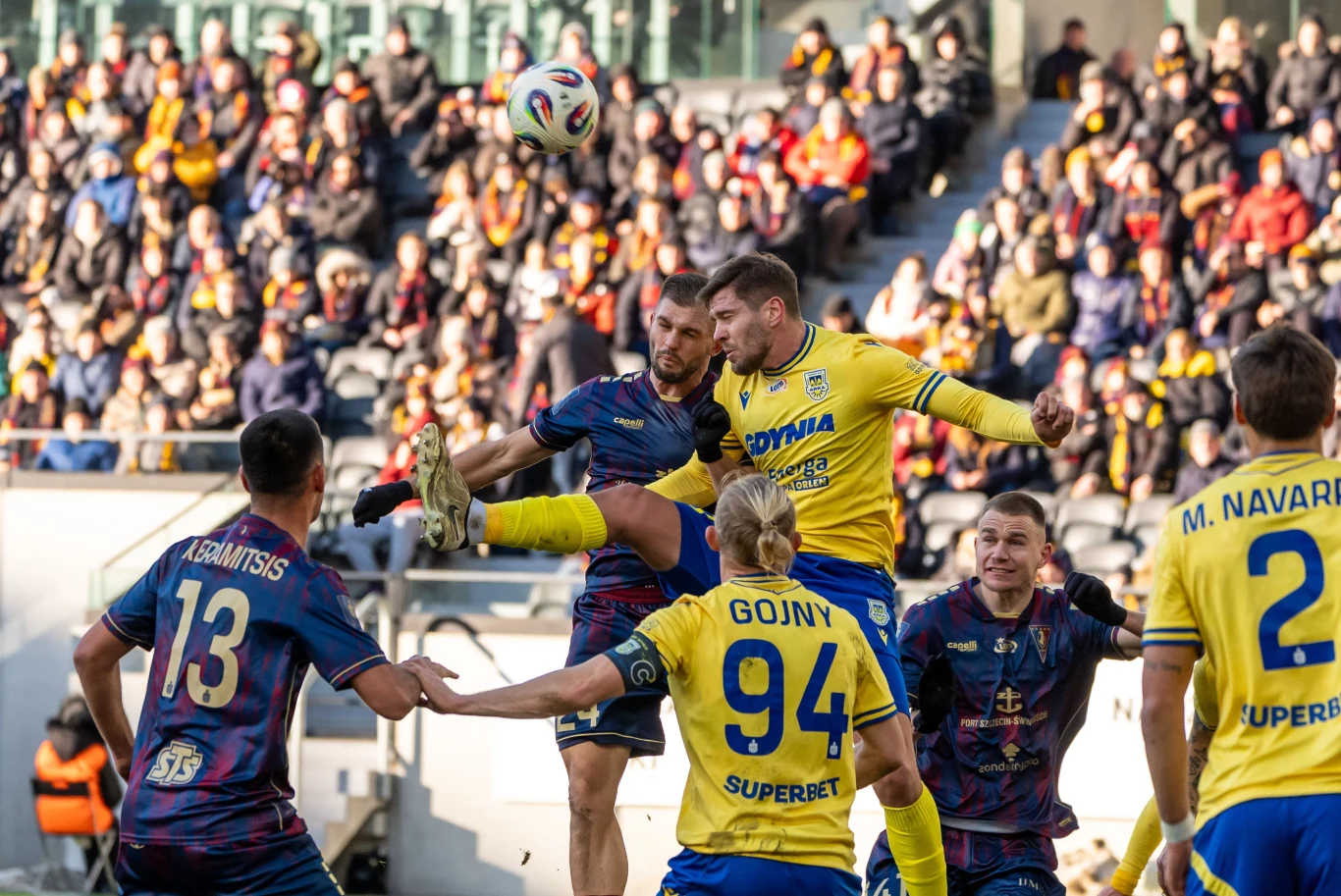 Arka Gdynia (żółte koszulki) pozostają jedynym zespołem, który w tym sezonie nie zgarnął jeszcze pełnej puli na wyjeździe Zawodnicy dwóch drużyn piłkarskich walczą o piłkę podczas meczu, w tle widoczna pełna trybuna kibiców, a główny pojedynek toczy się w powietrzu, gdzie piłkarz w żółtej koszulce próbuje strzelić głową.
