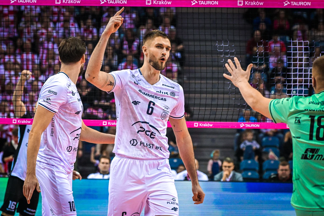 Siatkarz drużyny w białym stroju z numerem 6 na tle siatki sportowej unosi rękę w geście radości podczas meczu, wokół niego pozostali zawodnicy, a w tle widownia i banery reklamowe.