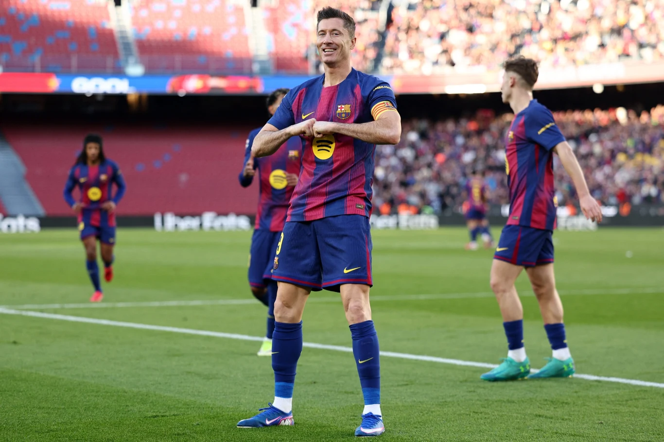 Robert Lewandowski Piłkarz FC Barcelony w granatowo-bordowej koszulce świętuje zdobycie bramki na murawie stadionu, wokół stoją inni zawodnicy drużyny, a w tle widoczna jest publiczność na trybunach.