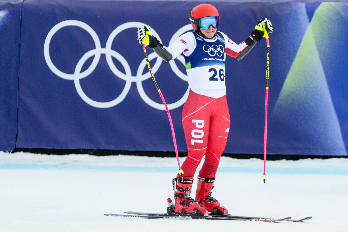 Zawodniczka w czerwonym stroju narciarskim z napisem POL, w kasku i goglach, trzymająca kijki narciarskie, znajduje się na stoku podczas zawodów olimpijskich, w tle widoczny symbol olimpijski.
