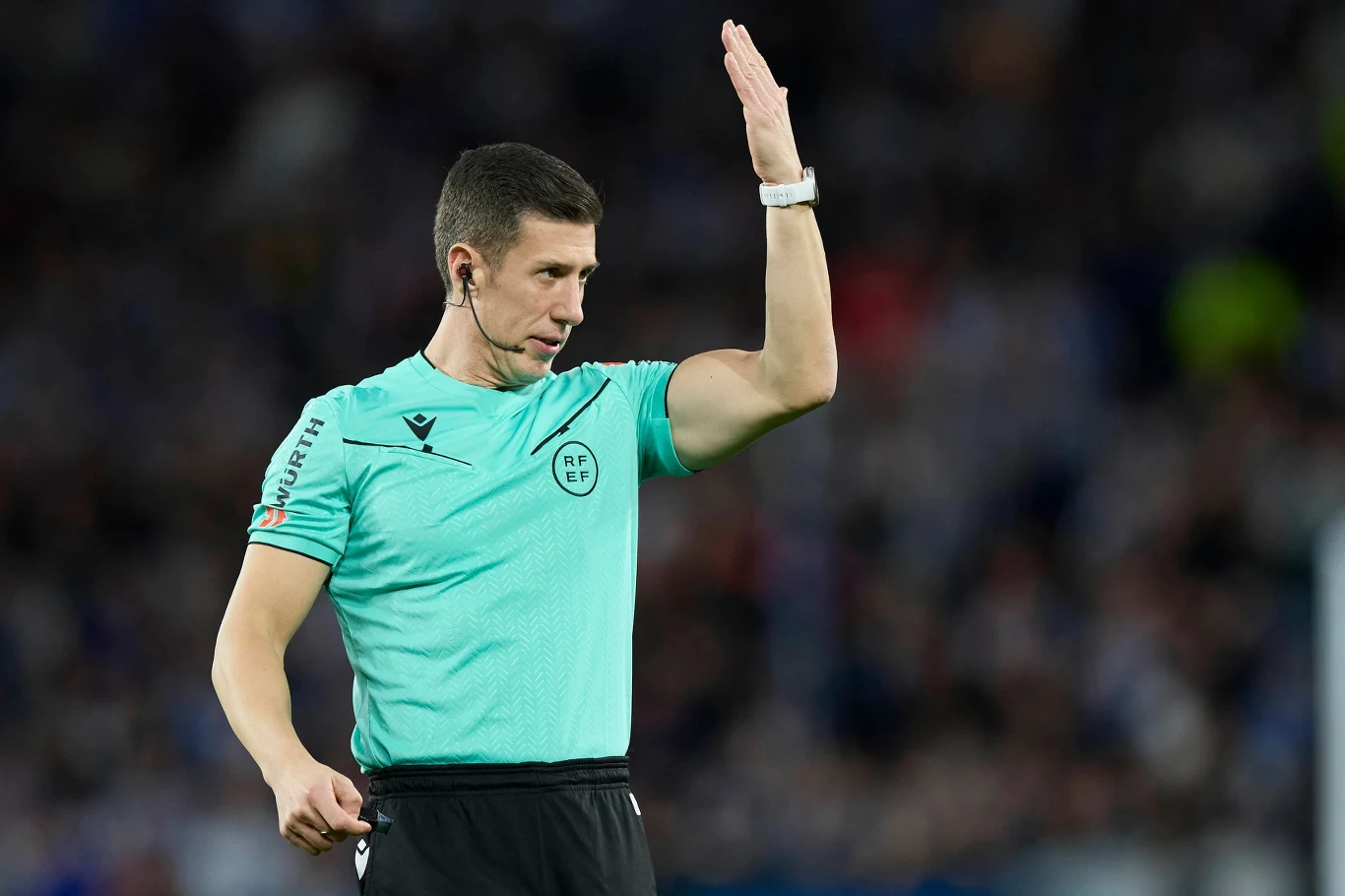 Sędzia piłkarski w zielonej koszulce sygnalizuje decyzję podczas meczu piłki nożnej, trzymając rękę uniesioną do góry na tle rozmytego stadionu i kibiców.