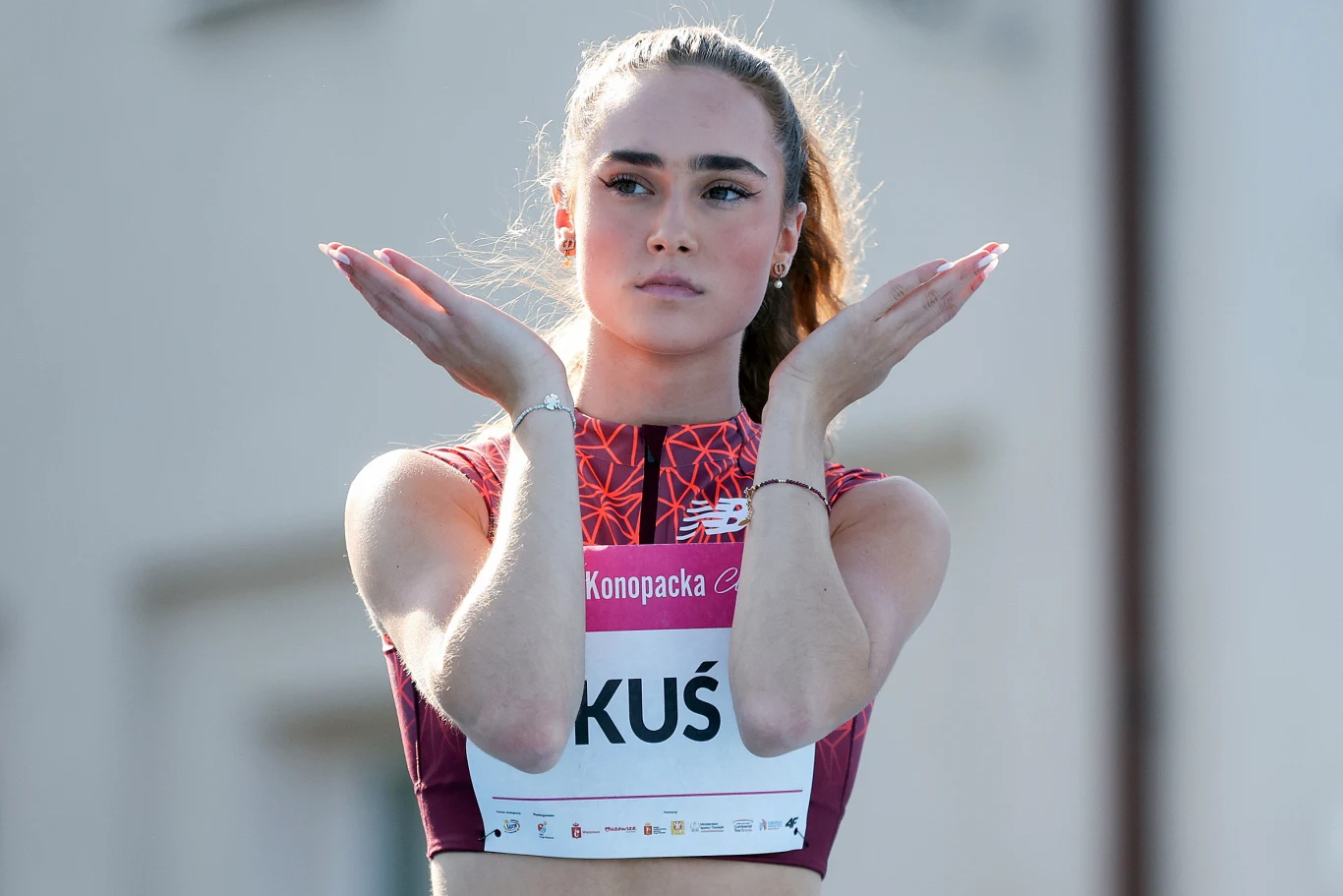 Anastazja Kuś Młoda sportsmenka w sportowym stroju wykonuje gest rękami przed klatką piersiową. Na piersi widoczny numer startowy z nazwiskiem KUŚ oraz oznaczenia sponsora. Tło rozmyte, skupienie na twarzy oraz sylwetce zawodniczki.