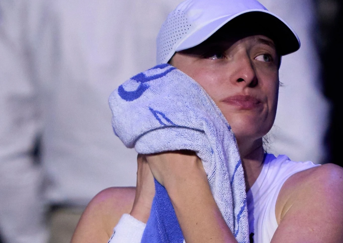 Kobieta w białej sportowej czapce i stroju trzyma przy twarzy niebiesko-biały ręcznik, z wyrazem silnych emocji oraz łez, na tle rozmytych postaci w tle.
