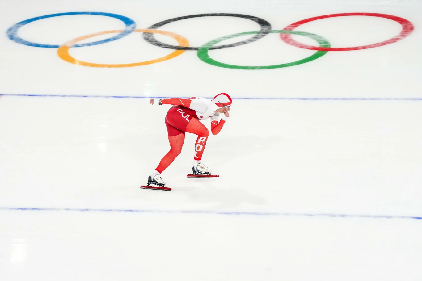 Łyżwiarz szybki w biało-czerwonym stroju, oznaczonym napisem 'POL' i narodowym godłem, porusza się po lodowej tafli, za nim duże kolorowe koła olimpijskie.