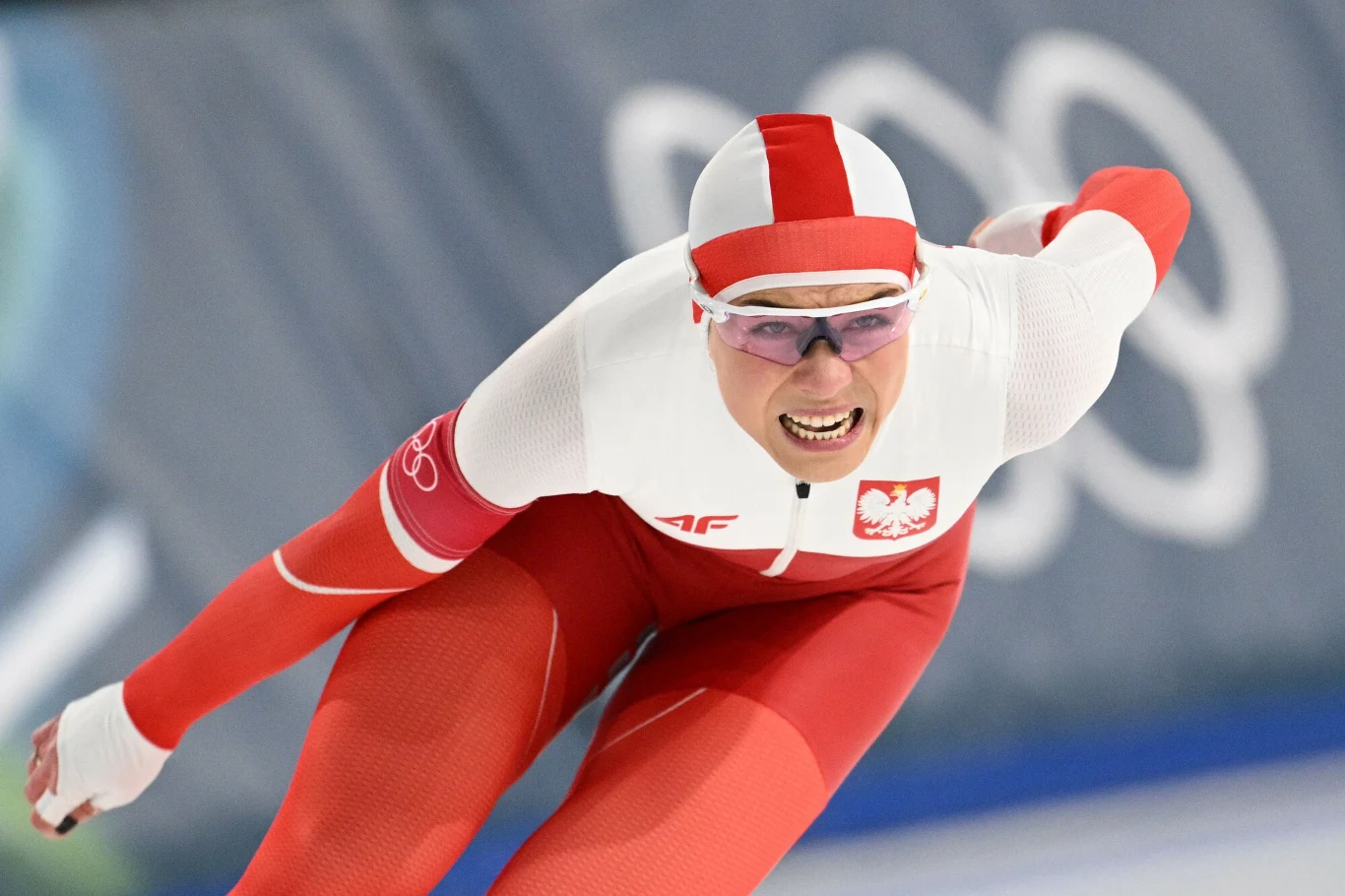 Kaja Ziomek-Nogal Zawodnik w biało-czerwonym stroju narodowym Polski podczas wyścigu na łyżwach startuje dynamicznie na tle olimpijskich kół.