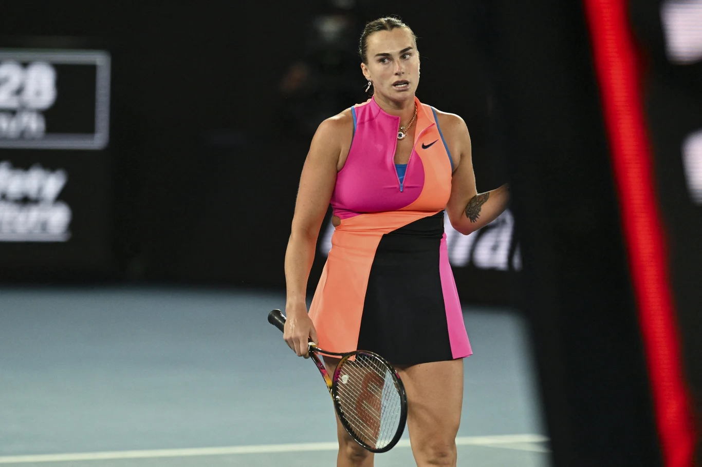 Zawodowa tenisistka w sportowej sukience w odcieniach różu, pomarańczu i czerni, trzymająca rakietę tenisową na korcie podczas meczu, w tle rozmazane trybuny i tablice świetlne.