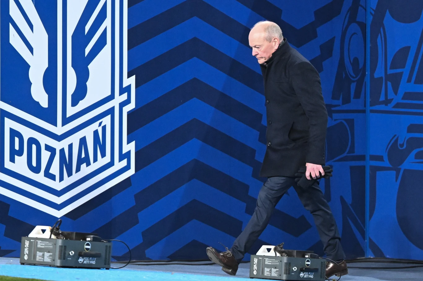 Starszy mężczyzna ubrany w ciemny płaszcz idzie obok dużego, niebieskiego baneru z napisem 'Poznań' oraz graficznym symbolem związanym z miastem lub klubem sportowym. Na ziemi stoją skrzynki techniczne.