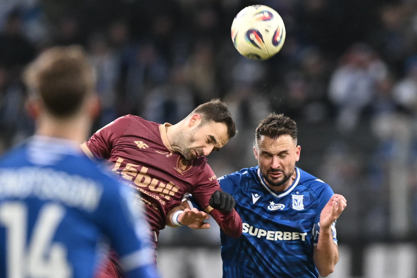 Dwóch piłkarzy rywalizuje o piłkę głową podczas meczu, jeden ubrany w bordowy strój, drugi w niebieski; wyraziste emocje na twarzach zawodników podkreślają intensywność walki.