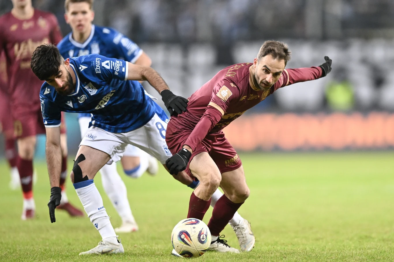 Dwóch piłkarzy walczących o piłkę podczas meczu piłki nożnej, jeden ubrany w granatowy strój, drugi w bordowy, dynamiczna akcja na murawie, widoczny wysiłek fizyczny i sportowa rywalizacja.