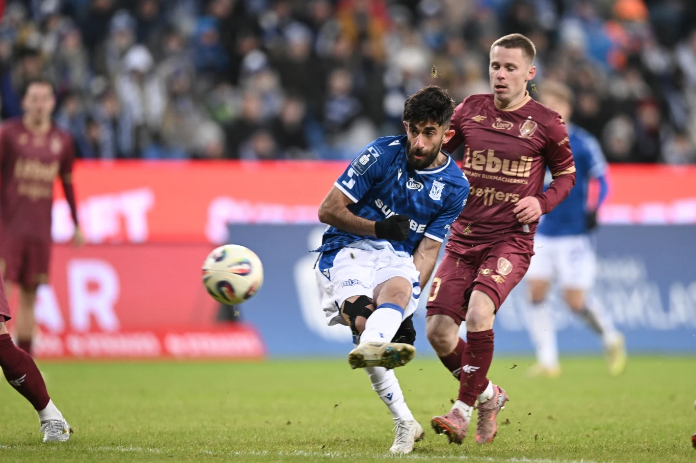 Dwóch piłkarzy w trakcie meczu piłkarskiego, zawodnik w niebieskiej koszulce oddaje strzał na bramkę, a zawodnik w bordowym stroju próbuje go zablokować. W tle widoczna publiczność na stadionie.