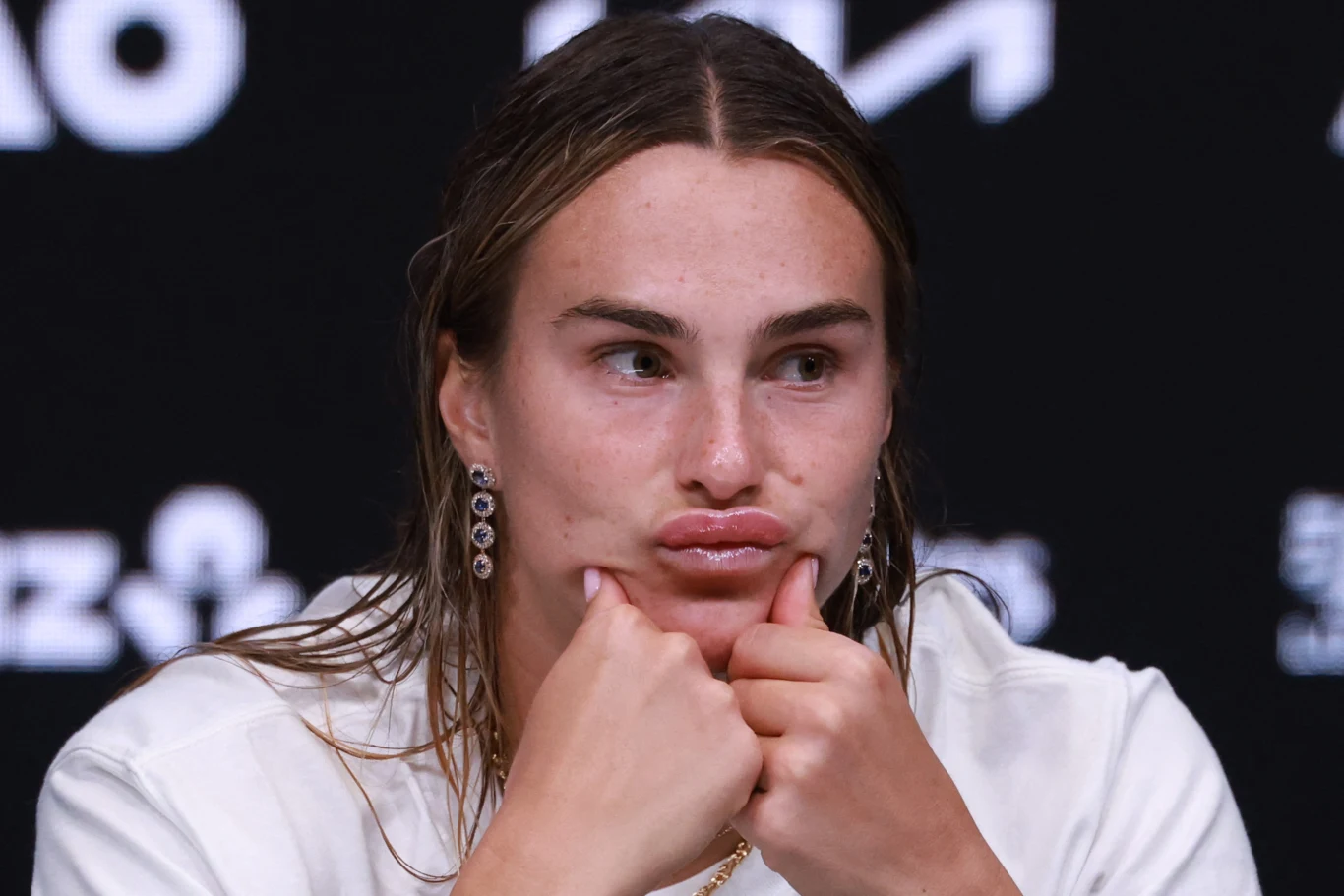 Aryna Sabalenka Młoda kobieta w białej koszulce siedzi z założonymi dłońmi na brodzie, robiąc zabawną minę, na jej uszach wiszą ozdobne kolczyki, w tle rozmazane logo i napisy.