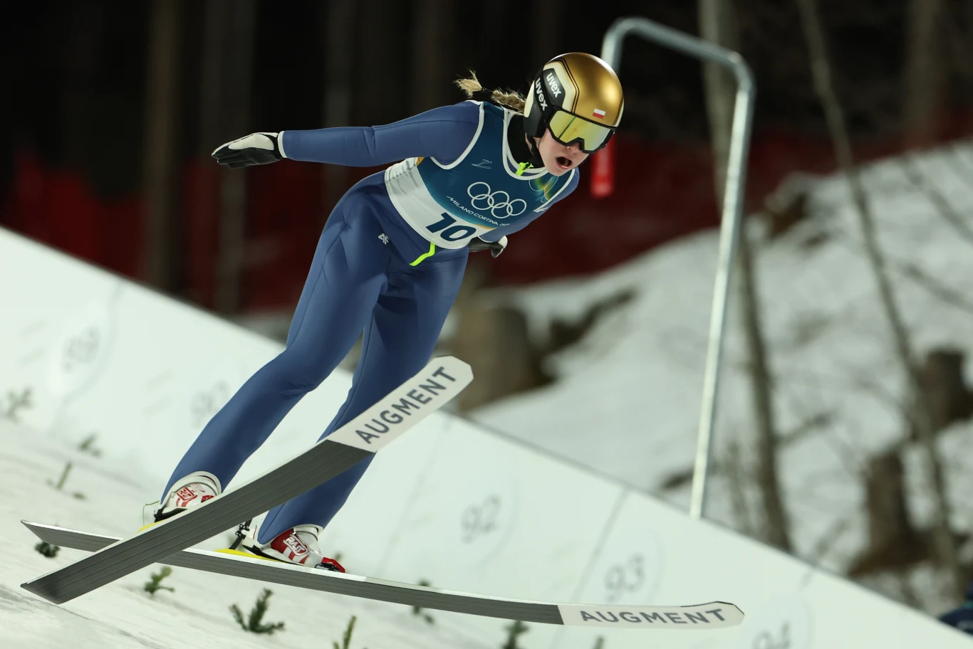 Skoczek narciarski w locie podczas zawodów, w niebieskim kombinezonie i złotym kasku, na tle śnieżnego zeskoku oraz olimpijskich oznaczeń.