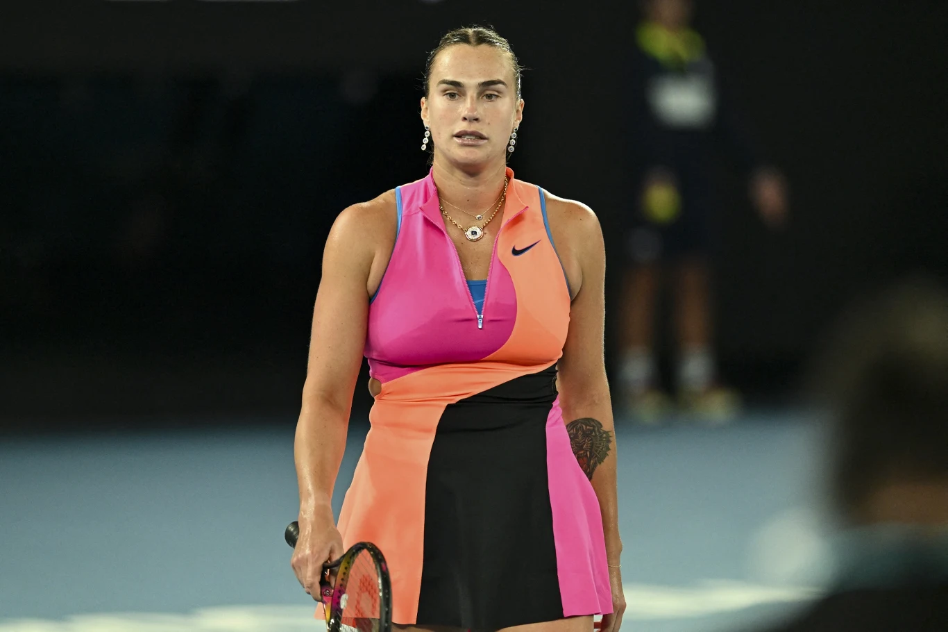 Zawodniczka tenisowa w kolorowej sukience sportowej trzymająca rakietę na korcie podczas meczu przy sztucznym oświetleniu, w tle niewyraźna sylwetka innej osoby.