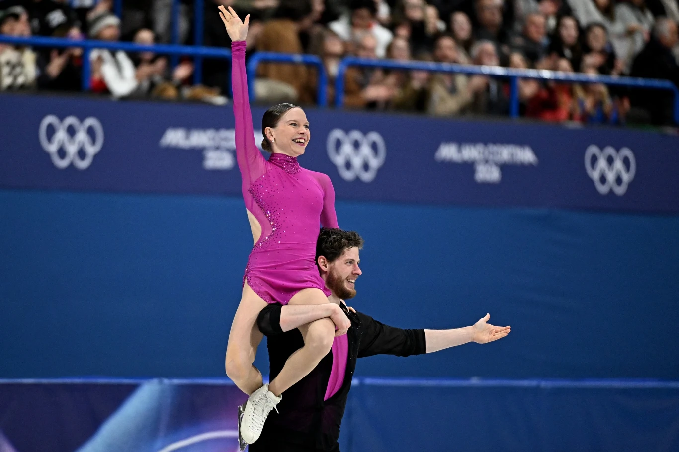 Para łyżwiarzy figurowych wykonuje widowiskową akrobację na lodzie, kobieta w różowym stroju jest trzymana przez partnera, wokół zgromadzona publiczność obserwuje występ podczas zawodów olimpijskich.