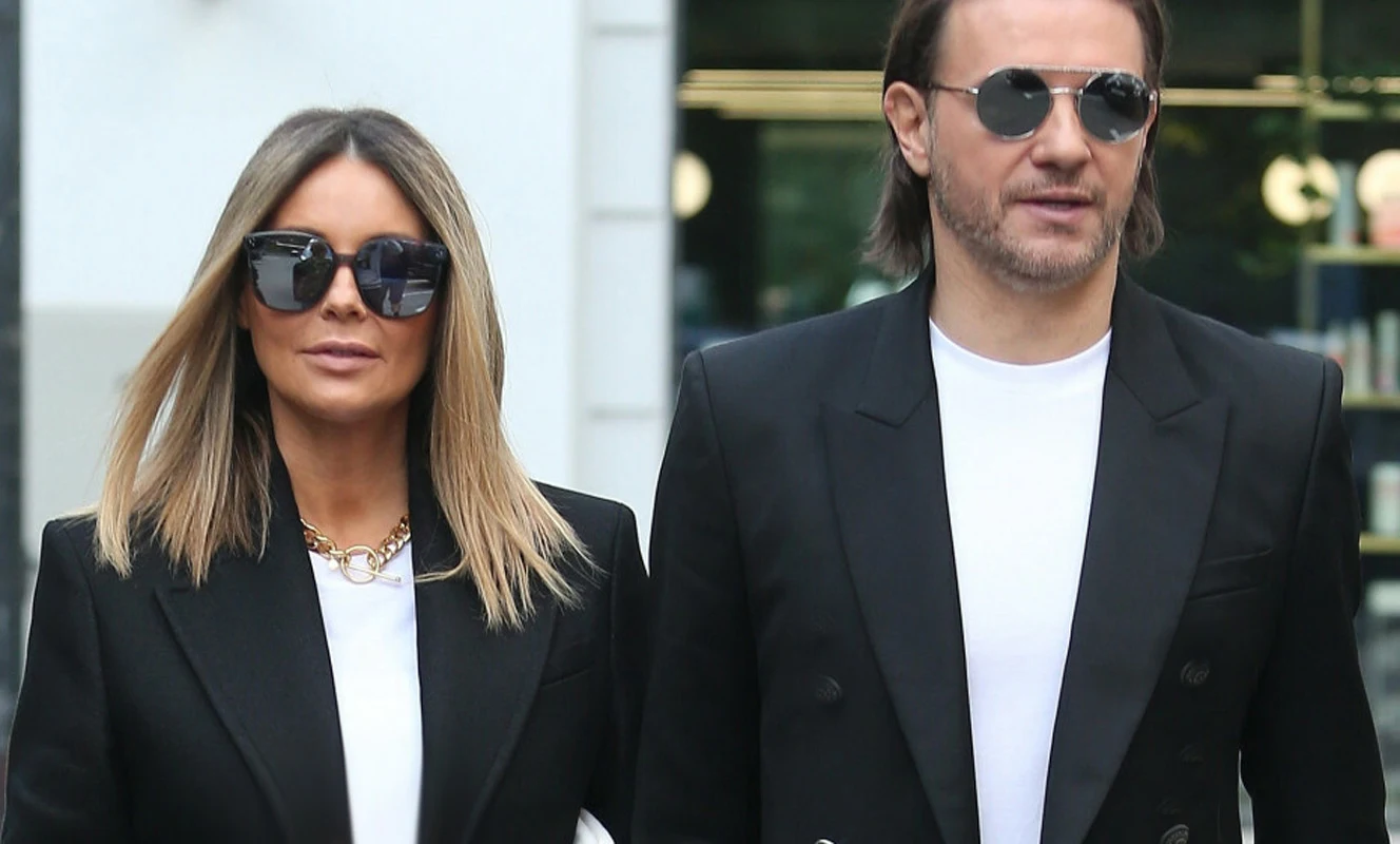 Rozenek i Majdan Para ubrana elegancko, oboje w ciemnych marynarkach i białych koszulkach, noszący ciemne okulary przeciwsłoneczne, z poważnymi wyrazami twarzy, spacerują razem w przestrzeni miejskiej.