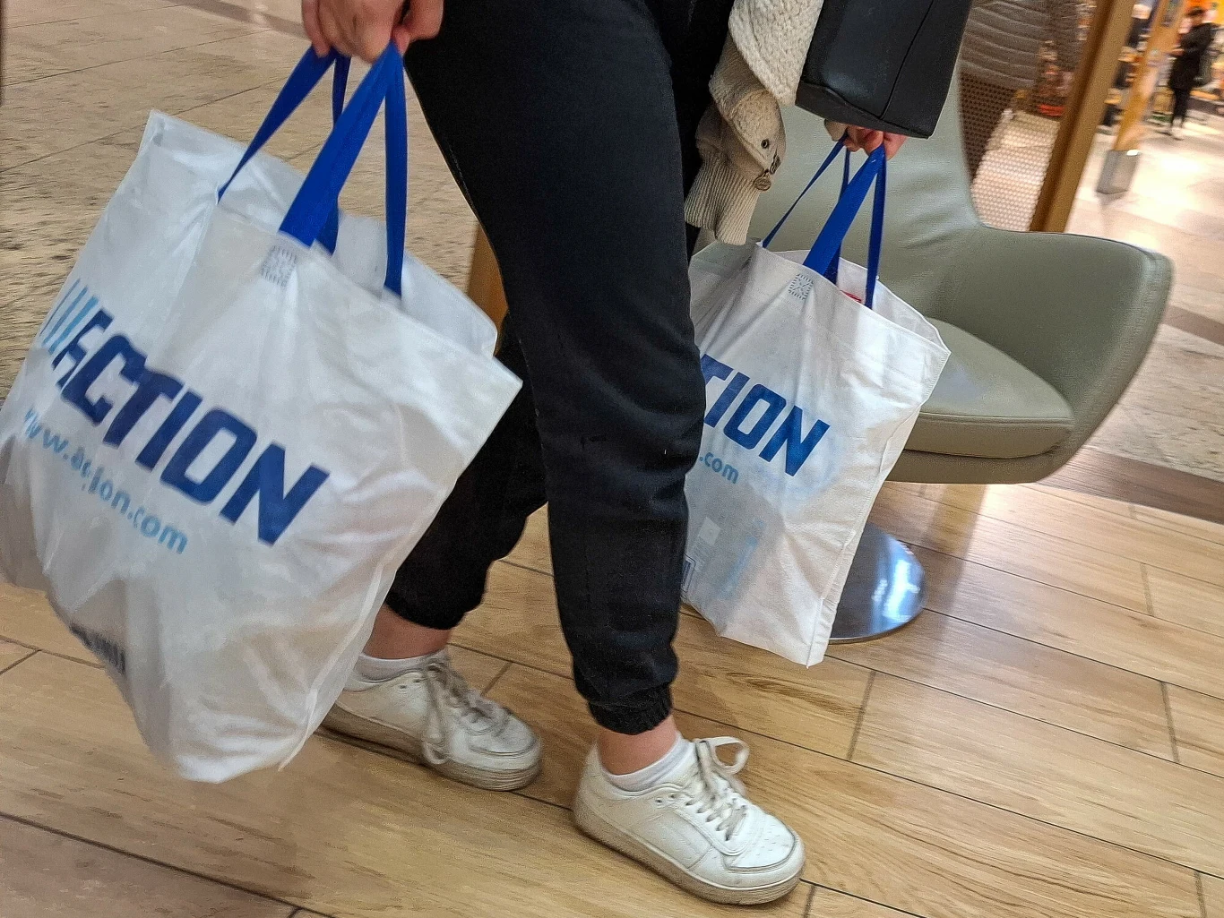 Sieć Action pilnie wycofuje jeden ze swoich produktów, ponieważ zawiera azbest Osoba ubrana w ciemne spodnie i białe buty sportowe niesie dwie torby zakupowe z niebieskimi uchwytami, idąc po jasnej, drewnianej podłodze w przestrzeni handlowej.