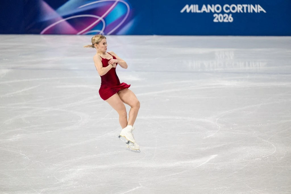 Łyżwiarka figurowa w czerwonej sukni wykonuje skok na lodowisku podczas zawodów olimpijskich Milano Cortina 2026. W tle widoczny baner z nazwą wydarzenia. Ujęcie dynamiczne, oddające ruch sportowca.