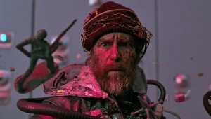 Mężczyzna z potarganą brodą i skupionym wyrazem twarzy, ubrany w metaliczną kurtkę oraz czapkę z przyszytymi kablami i elementami elektroniki, otoczony latającymi przedmiotami, w tym figurką żołnierza oraz przewodami.