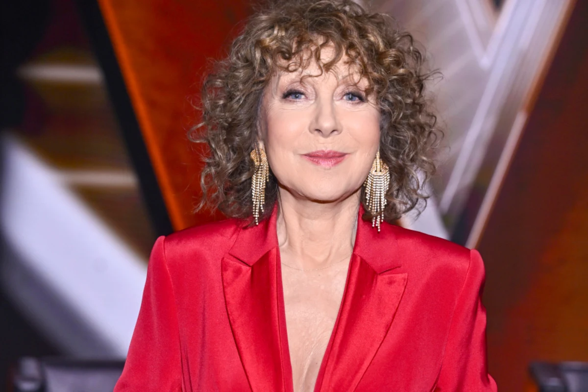 Alicja Majewska Kobieta o kręconych, jasnobrązowych włosach ubrana w elegancką, czerwoną marynarkę, z długimi złotymi kolczykami, uśmiechnięta, w tle rozmyte elementy scenografii.