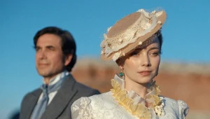 Kobieta w eleganckim, kremowym stroju z epoki z ozdobnym kapeluszem i żółtymi kwiatami przy szyi stoi na pierwszym planie, za nią widoczny mężczyzna w garniturze z apaszką wokół szyi, oboje na tle błękitnego nieba.