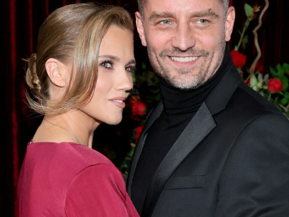 Agnieszka Kaczorowska, Marcin Rogacewicz Elegancka kobieta w czerwonej sukni i mężczyzna w czarnym garniturze stoją razem, uśmiechnięci, na tle czerwonych zasłon i kwiatów, pozują do wspólnego zdjęcia.