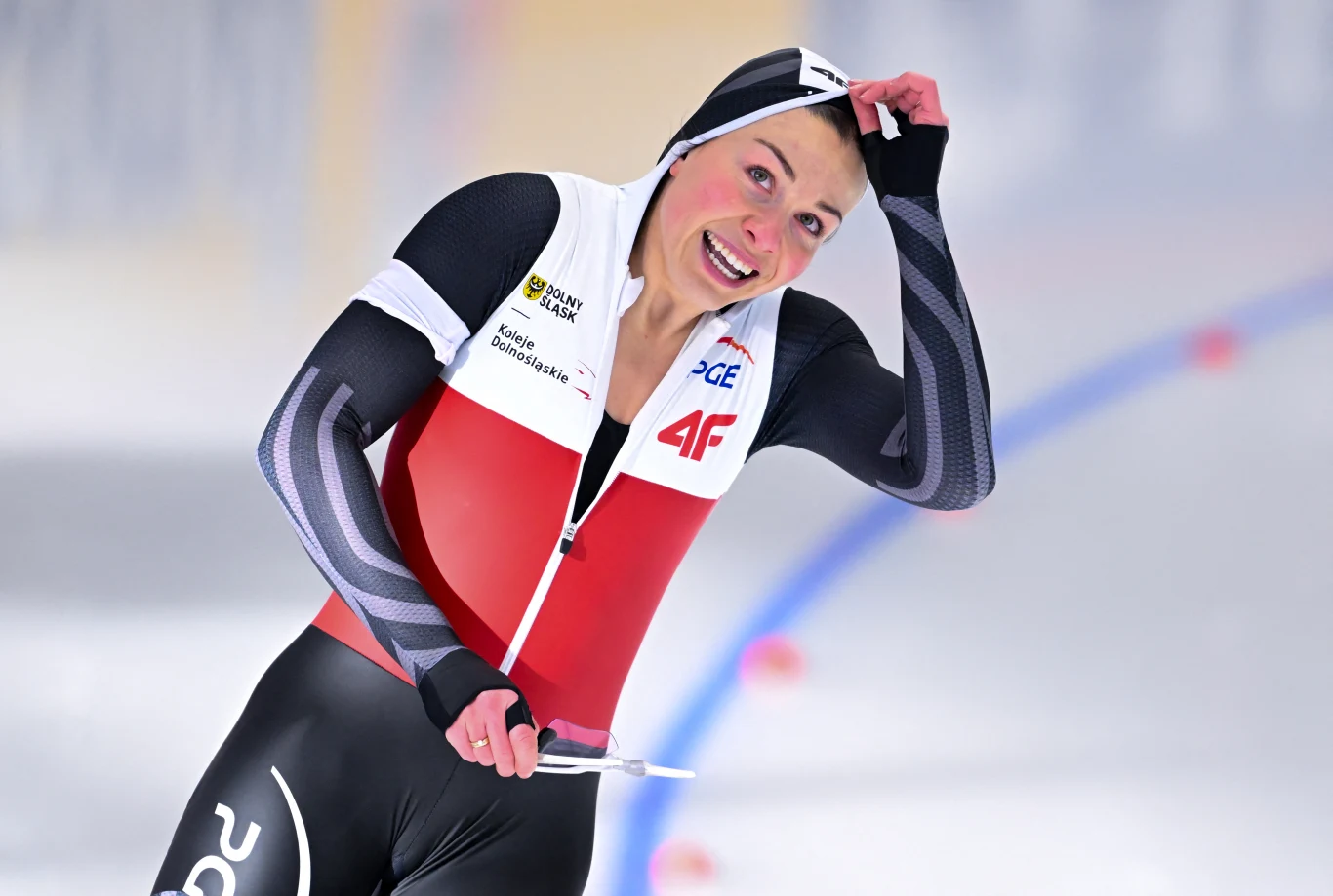 Zawodniczka w stroju panczenisty na torze lodowym po ukończeniu wyścigu z uśmiechem na twarzy, unosząca rękę do głowy. Na stroju widoczne logotypy sponsorów, tło rozmyte z widocznymi nieostrymi liniami toru.