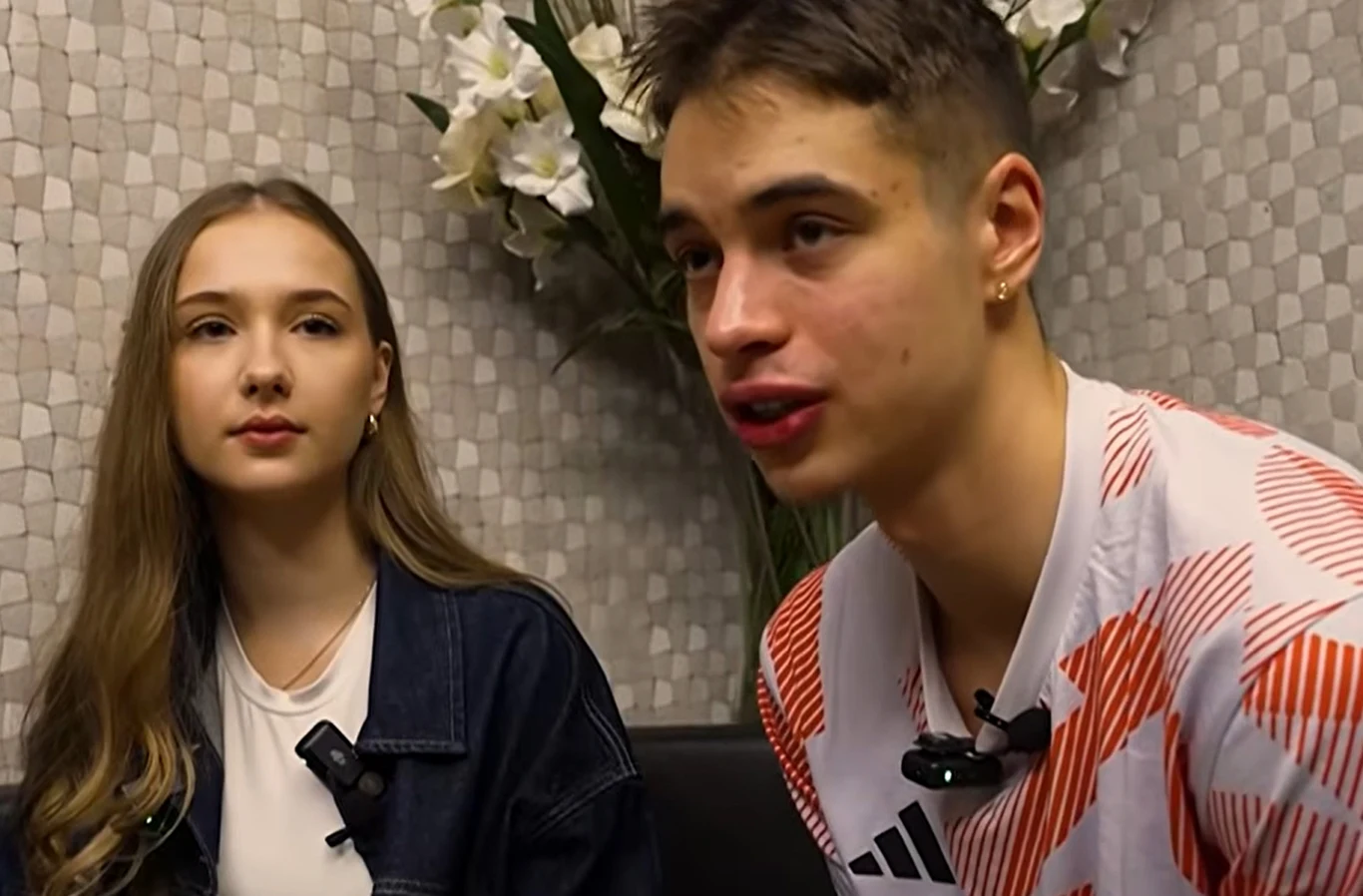 Władimir Semirunnij ze swoją dziewczyną Klaudią Młoda kobieta i młody mężczyzna siedzą razem w pomieszczeniu, oboje mają mikrofony przypięte do ubrań, w tle widoczne są kwiaty i jasna tapeta.