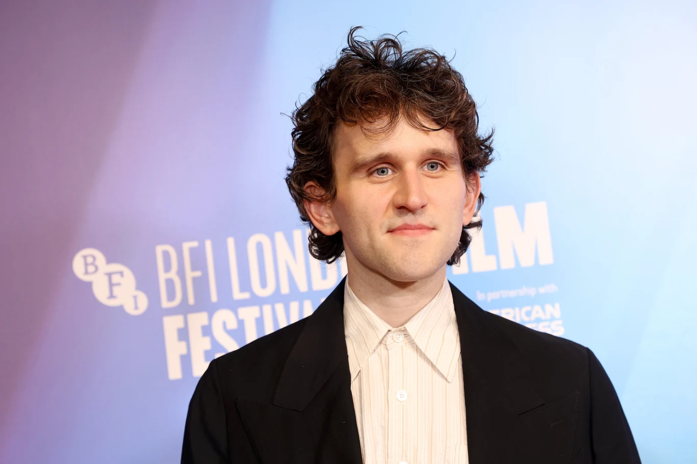 Harry Melling Mężczyzna w marynarce i jasnej koszuli stoi na tle niebieskiej ścianki z logotypami festiwalu filmowego BFI London Film Festival.