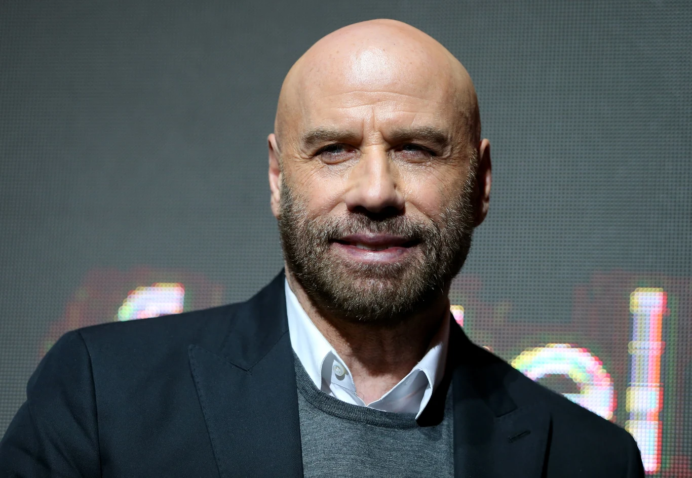 John Travolta Elegancko ubrany, łysy mężczyzna z brodą, uśmiechnięty, stoi na tle rozmytego napisu lub grafiki, światło skupione na jego twarzy.