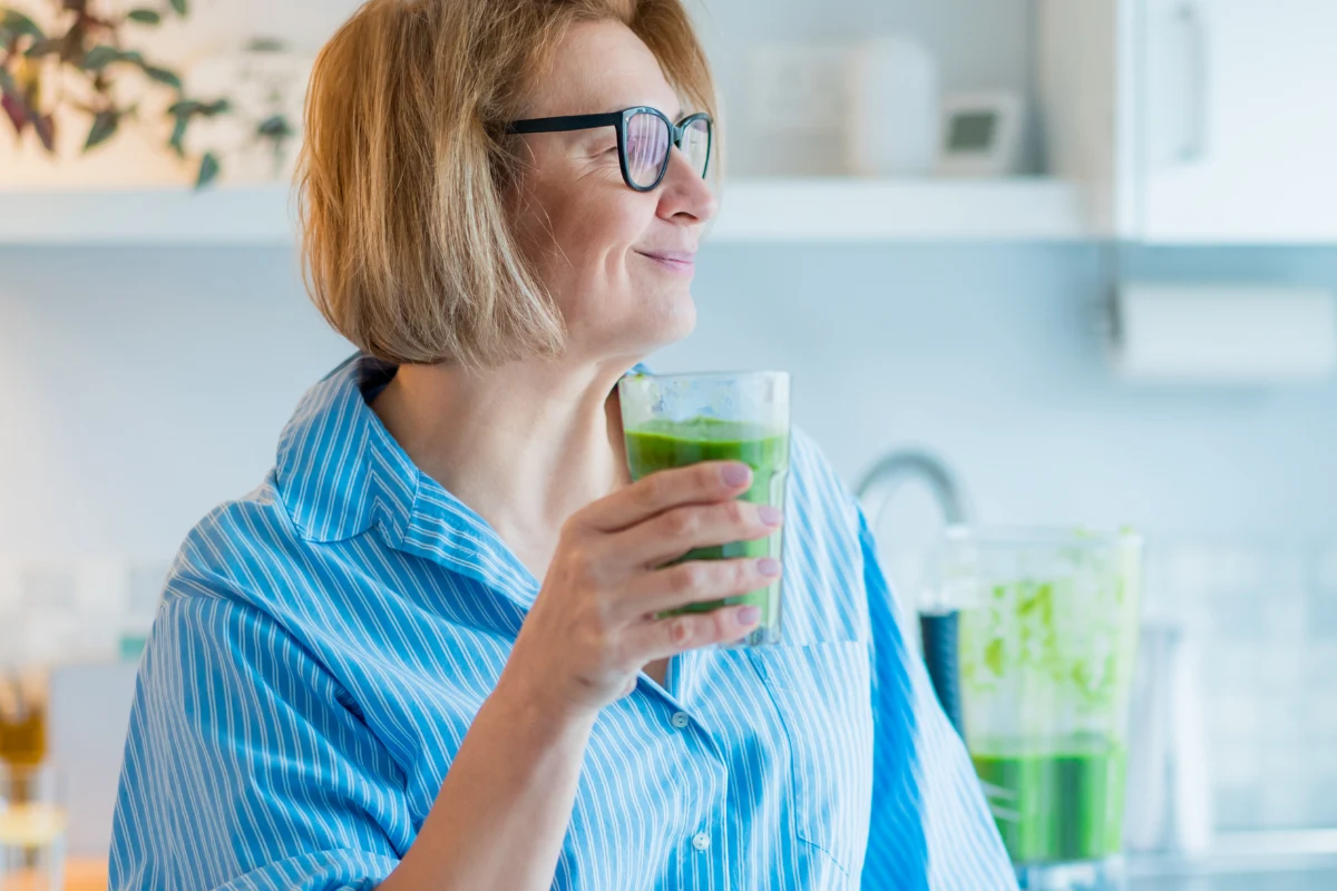 Łyżeczka moringi do koktajlu to dziś jeden z najpopularniejszych sposobów na naturalne wsparcie metabolizmu Kobieta w okularach i niebieskiej koszuli pije zielony koktajl w jasnej kuchni. Na blacie w tle stoi blender z resztką zielonego napoju.