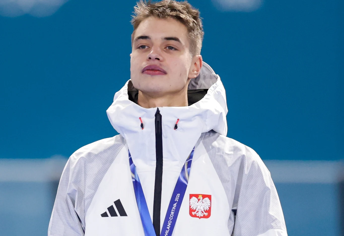 Władimir Semirunnij, zimowe igrzyska olimpijskie Mediolan-Cortina 2026 Młody mężczyzna w sportowej kurtce z orłem na piersi i logo Adidas, z medalem na szyi, stoi na tle niebieskiego rozmytego tła.